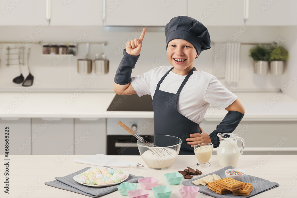 The boy in the cook's uniform smiles and holds his finger up. Preparation of cream, ice cream. Modern white kitchen. Dessert, chocolate,milk, towels, silicone molds whisk The boy in the cook's uniform smiles and holds his finger up. Preparation of cream, ice cream. Modern white kitchen. Dessert, chocolate,milk, towels, silicone molds whisk