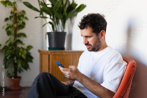 White adult man sitting on a living room with plants and white background while using smartphone. People using smartphones concept with copyspace.