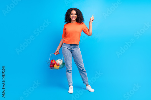 Full size photo of young afro woman happy smile hold basket shop point finger empty space ad promo isolated over blue color background