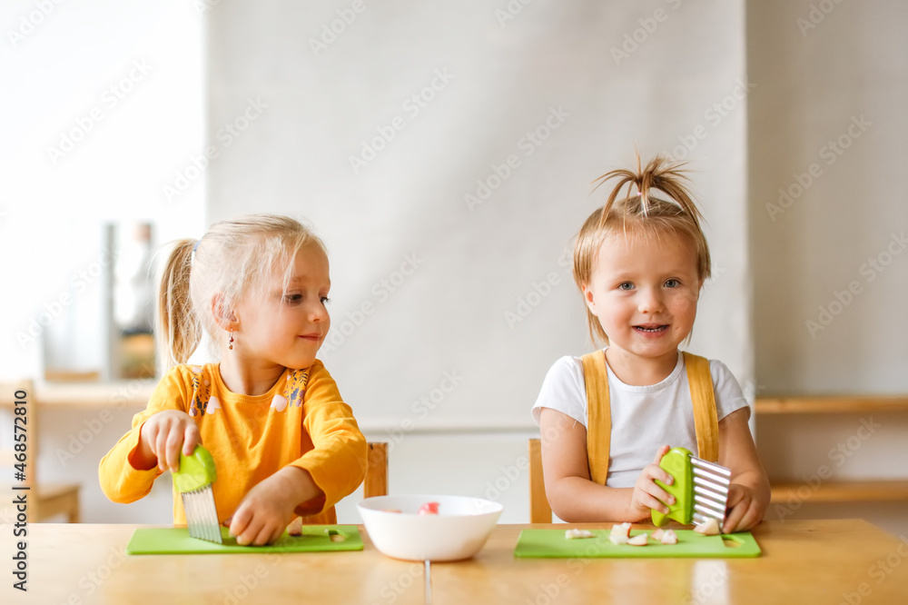 Cute kids toddlers girls at the table cut apples with safe knives, children cut food themselves, montessori and independence in kindergarten