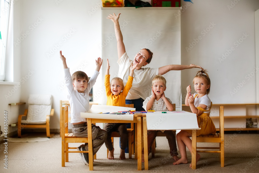 cheerful teacher woman with a group of cute preschool children at the table draw on paper with multicolored felt-tip pens, montessori and creativity, creative development and lesson in kindergarten - obrazy, fototapety, plakaty 