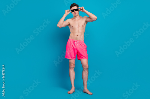 Photo of cool self-assured guy posing barefoot wear sunglass swimwear shorts isolated blue color background