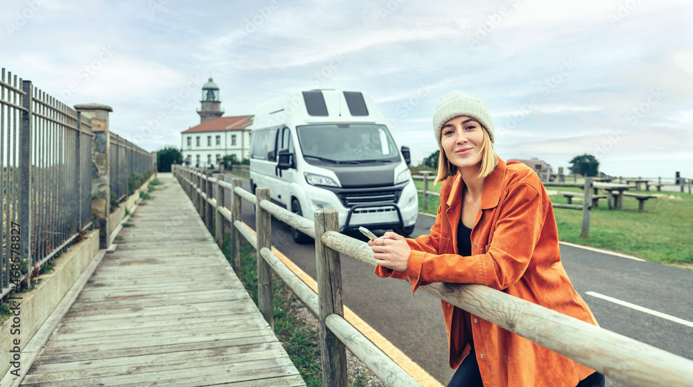 Young woman looking camera leaning on fence in front of camper van with lighthouse in background Young woman looking camera leaning on fence in front of camper van with lighthouse in background