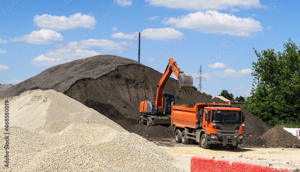 a crawler excavator loads crushed stone and sand into a truck. Large hills and piles of sand, gravel, crushed stone of white, gray and black color. a crawler excavator loads crushed stone and sand into a truck. Large hills and piles of sand, gravel, crushed stone of white, gray and black color.