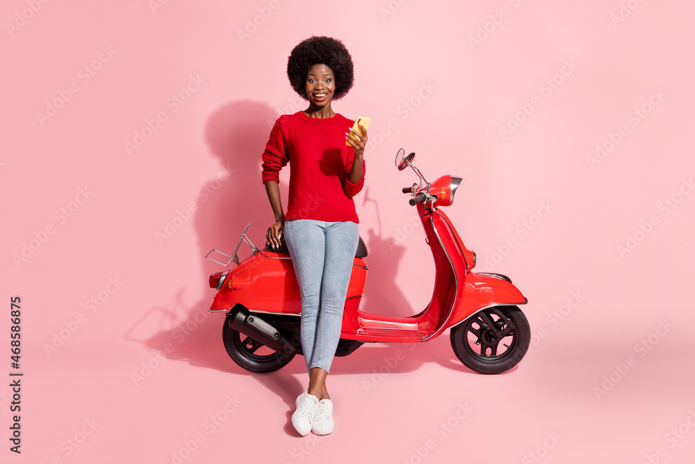 Full body photo brunette afro american girl wear casual outfit hold phone sit motorbike excited isolated on pink color background Full body photo brunette afro american girl wear casual outfit hold phone sit motorbike excited isolated on pink color background