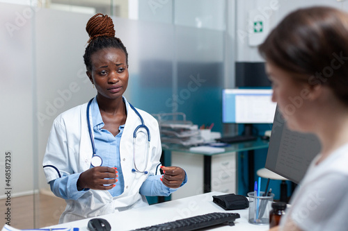 African american therapist doctor discussing with patient monitoring disease symptoms during medical examination in hospital office. Practitioner woman in uniform explaining medication treatment