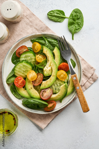 Avocado, red, yellow, black cherry tomato, spinach and cucumber fresh salad with spices pepper and olive oil in grey bowl on grey slate, stone or concrete background. Healthy food concept. Top view.