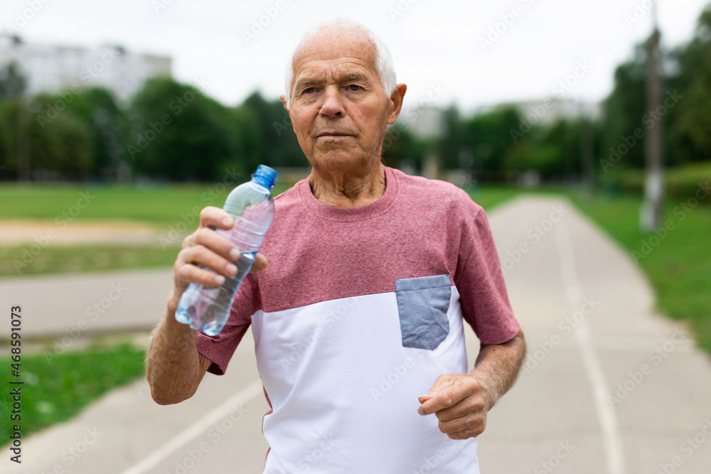 Senior man resting and drinking water from bottle after jogging in park Senior man resting and drinking water from bottle after jogging in park