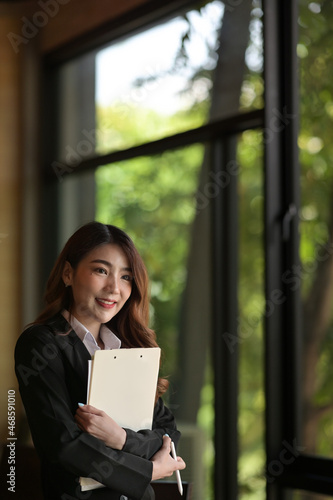Photo of a young businesswoman standing and holding a document and clipboard over a comfortable meeting room as a background.