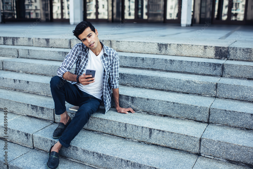 Portrait of young hipster guy in casual clothes holding modern cellular device and looking at camera while resting at urban stairs, Caucaisan man with smartphone posing during travel leisure in city