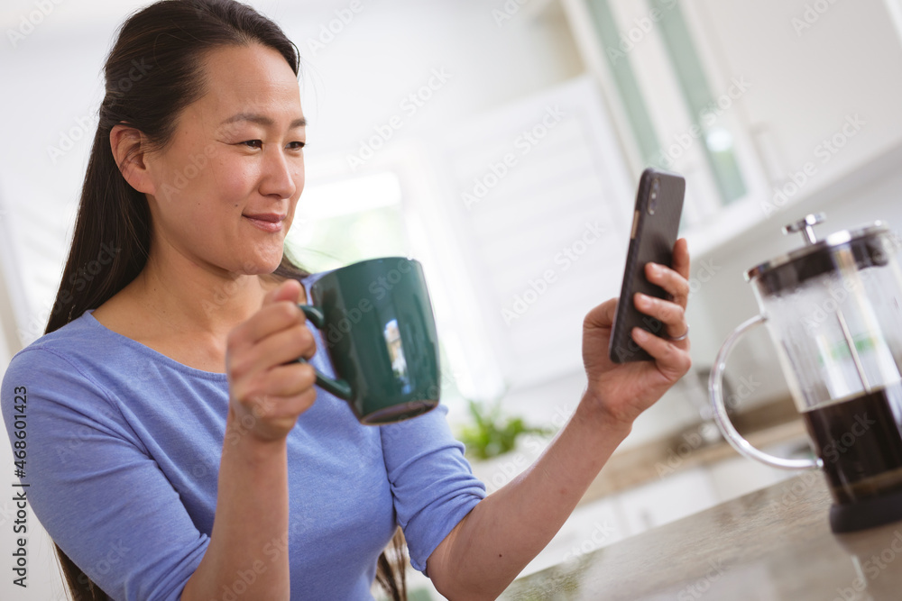 Happy asian woman drinking coffee and using smartphone in kitchen Happy asian woman drinking coffee and using smartphone in kitchen