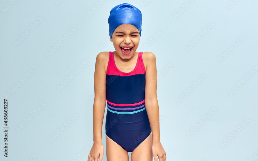 Studio image of a little funny happy girl in a swimsuit, isolated over blue studio background. A joyful wet kid in a swimsuit and swim cap smiling broadly during swimming training. Studio image of a little funny happy girl in a swimsuit, isolated over blue studio background. A joyful wet kid in a swimsuit and swim cap smiling broadly during swimming training.