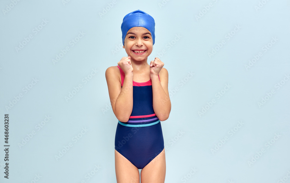 Studio image of a little funny happy girl in a swimsuit, isolated over blue studio background. A positive kid in swimwear making joyful gestures during wins swimming contest. Studio image of a little funny happy girl in a swimsuit, isolated over blue studio background. A positive kid in swimwear making joyful gestures during wins swimming contest.