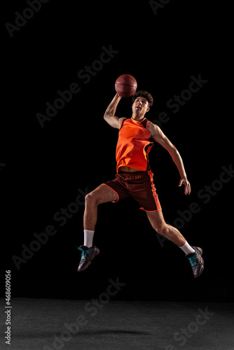 Full length portrait of basketball player training isolated on dark studio background. Tall muscular athlete jumping with ball. © master1305