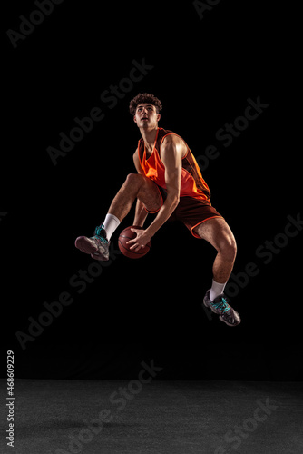 Full length portrait of basketball player training isolated on dark studio background. Tall muscular athlete jumping with ball.
