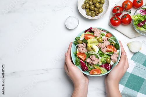Woman holding bowl of delicious salad with canned tuna at white table, top view. Space for text photo
