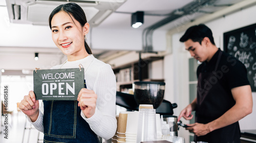 Young Asian female barista cafe owner holding the "WELCOME", "OPEN" announcement board with blur male barista in the background. Business with a partner. Successful entrepreneur teamwork.
