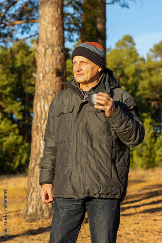 An elderly man drinks hot tea from a thermos on the shore of a lake with colorful autumn nature in the background. Hot coffee drink in nature.