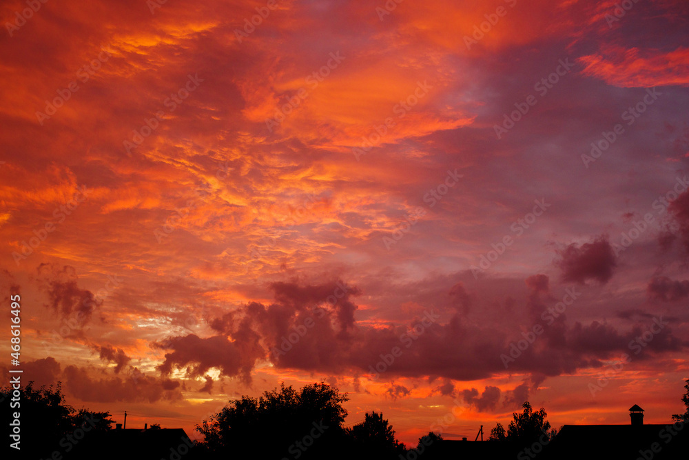 Red and orange cloudy sky before sunrise against a black silhos of rustic roofs and trees. August Landscape with Dawn Sky
