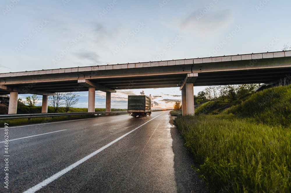 Beautiful nature landscape with a truck on a wet after rain asphalt road with a concrete bridge against the backdrop of a sunset and a beautiful sky with clouds.