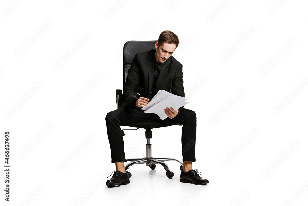 Portrait of young man in black business suit, office manager sitting on chair isolated on white background. Art, inspiration concept. Portrait of young man in black business suit, office manager sitting on chair isolated on white background. Art, inspiration concept.