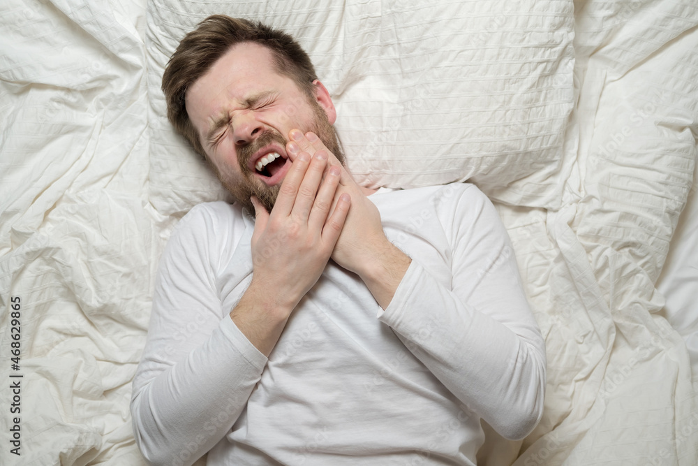 Man suffers from unbearable toothache, he holds hands on cheek and groans in pain, closing eyes and lying in bed. Top view. Close-up. Man suffers from unbearable toothache, he holds hands on cheek and groans in pain, closing eyes and lying in bed. Top view. Close-up.