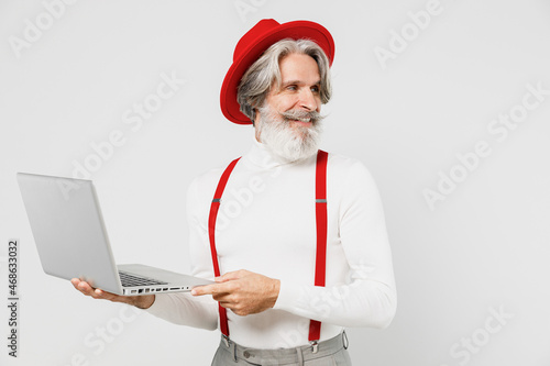 Elderly gray-haired mustache bearded man 60s in turtleneck red hat suspenders hold use work on laptop pc computer look aside isolated on plain white background studio portrait People lifestyle concept