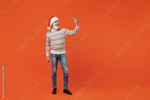 Full size body length old swanky bearded Santa Claus man 50s wears Christmas hat sweater doing selfie shot on mobile cell phone post photo on social network isolated on plain orange background studio.