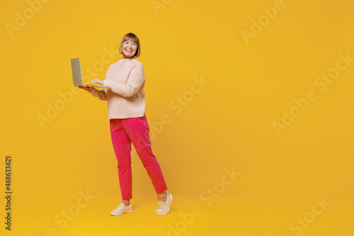 Full body elderly smiling happy caucasian copywriter woman 50s wears pink casual knitted sweater hold use work on laptop pc computer look aside isolated on plain yellow background studio portrait