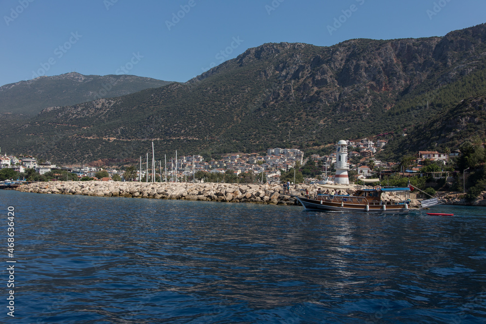 A lighthouse on top of the rocks away from the shore while a wooden boat passing by in front of it and big green mountains in the background and city of Kas on valley.