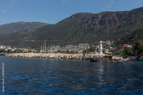 A lighthouse on top of the rocks away from the shore while a wooden boat passing by in front of it and big green mountains in the background and city of Kas on valley. © Arzi