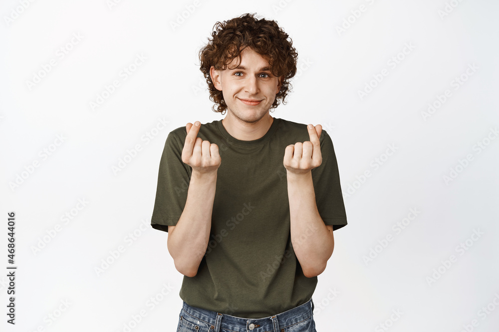 Cute curly-haired guy showing finger hearts gesture and smiling tenderly at camera. Romantic man making confession in love, standing over white background Cute curly-haired guy showing finger hearts gesture and smiling tenderly at camera. Romantic man making confession in love, standing over white background