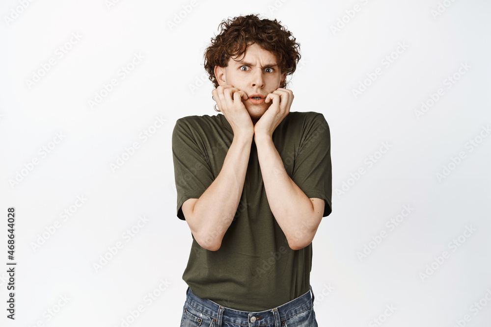 Scared young curly man gasping, trembling from fear, staring alarmed at camera, standing over white background in t-shirt Scared young curly man gasping, trembling from fear, staring alarmed at camera, standing over white background in t-shirt