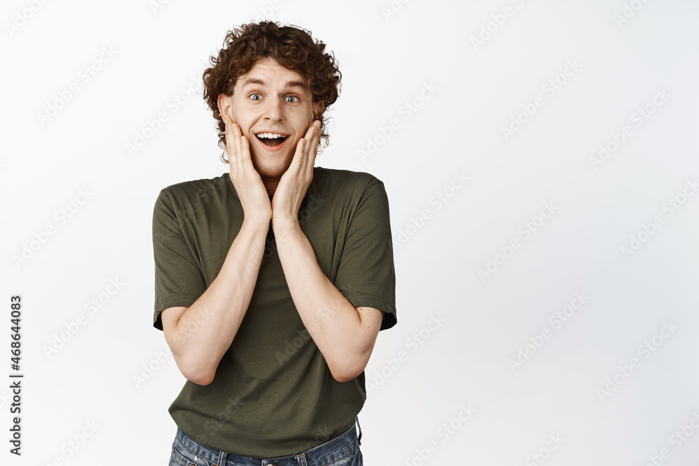 Happy handsome curly-haired man, cupping his face in hands, smiling and gazing fascinated at camera, standing over white background Happy handsome curly-haired man, cupping his face in hands, smiling and gazing fascinated at camera, standing over white background