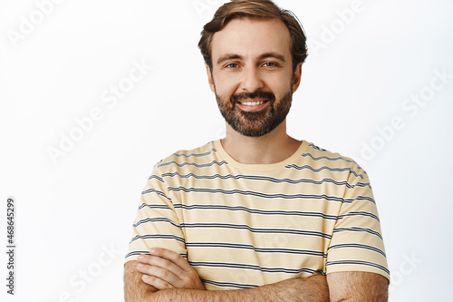 Close up portrait of handsome man with beard, smiling and looking with confident face, cross arms like professional, standing over white background