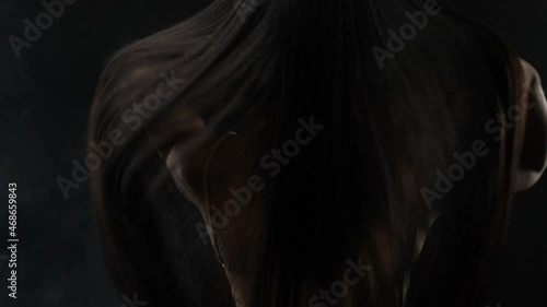 Close-up of a brunette with luxurious hair on a dark background. The girl stands with her back and develops her hair. photo