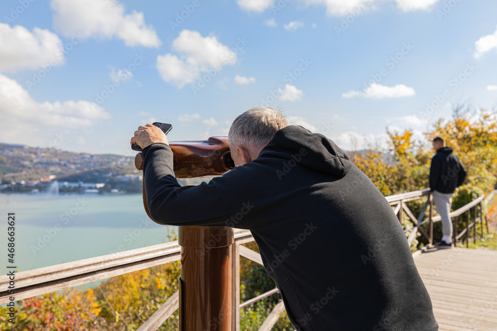 A man looks through a tourist telescope at the lake and mountains. Autumn landscape.
