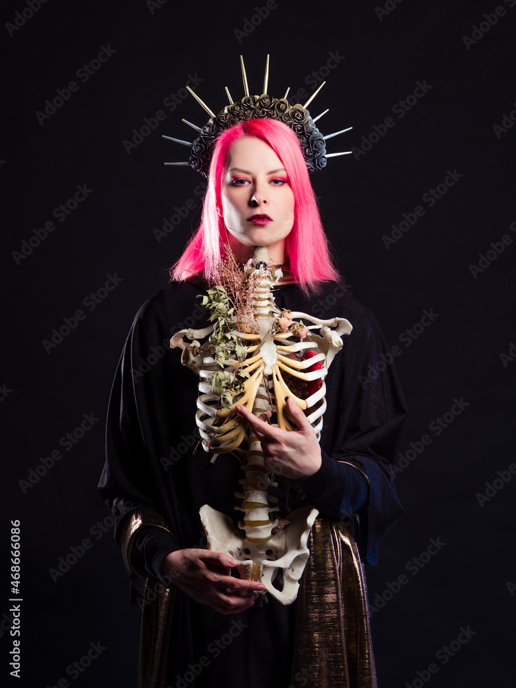 Naklejka premium A Gothic queen in a crown with roses and rays holds a skeleton of a man without a head in her arms. Young woman with pink hair in a mystical image, portrait on black