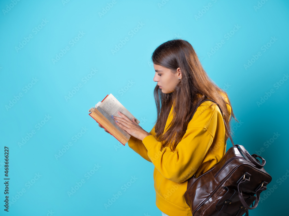 A student with a book in her hands, reading a textbook and preparing for a new lesson. A young brunette in a yellow hoodie and with a backpack, leafing through a book. A student with a book in her hands, reading a textbook and preparing for a new lesson. A young brunette in a yellow hoodie and with a backpack, leafing through a book.