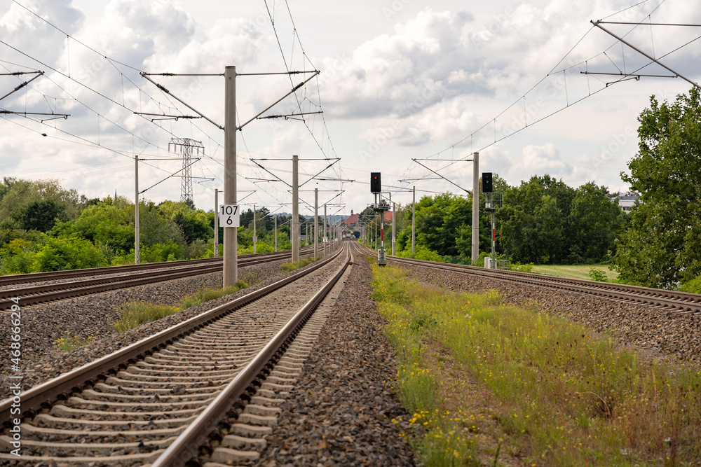 Railroad tracks in the green nature. Steel tracks, light signs and electric poles. Beautiful white clouds on the overcast sky on a summer day. Empty railways heading to a small village.