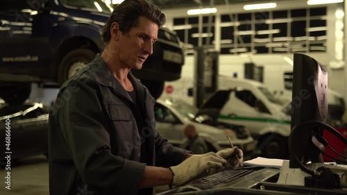 Serious atmosphere. Waist up portrait view of the auto mechanic standing near his computer and looking at the screen with car at the background. Car repair concept