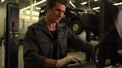 Waist up portrait view of the auto mechanic standing near his computer and checking orders with opened car at the background. Car repair concept