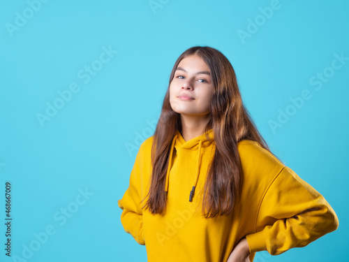 A beautiful young brunette in a yellow oversize hoodie, a positive student in a bright outfit, portrait on a blue background