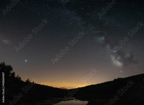 Milky Way reflected in a lake on the top of the mountain and with the lights of Portugal behind