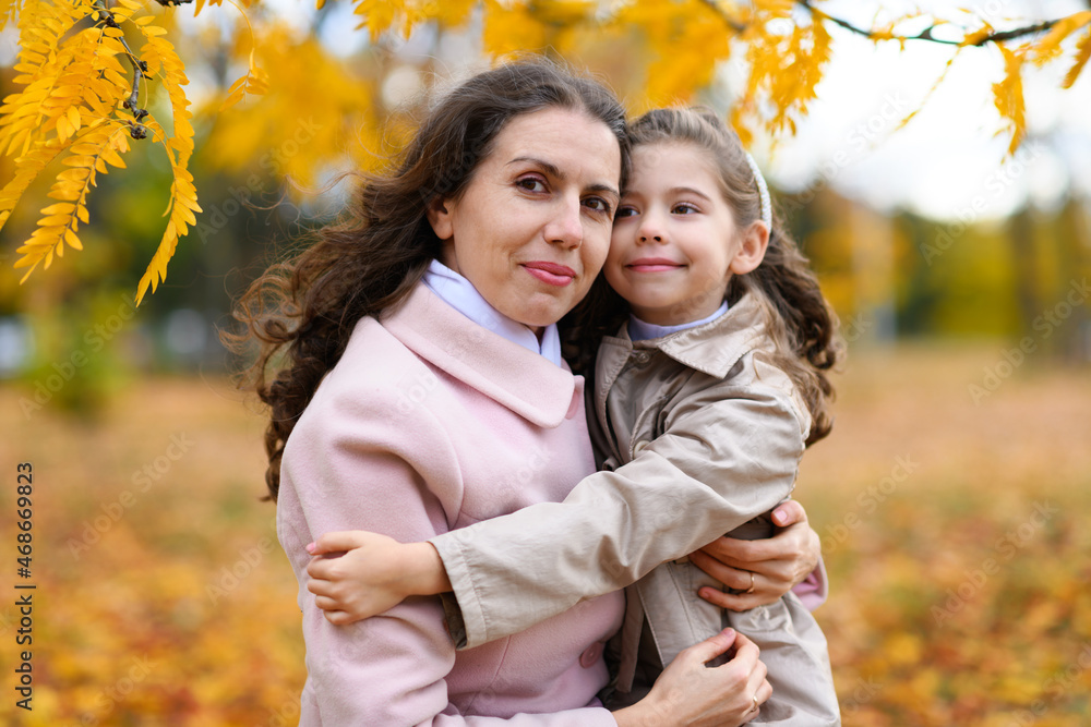 Mother and daughter portrait in an autumn park. Happy people pose against the background of beautiful yellow trees. They hug and are happy together.