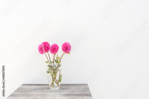 Glass vase with bright pink dahlia flowers on rustic wooden table against textured white wall with copy space