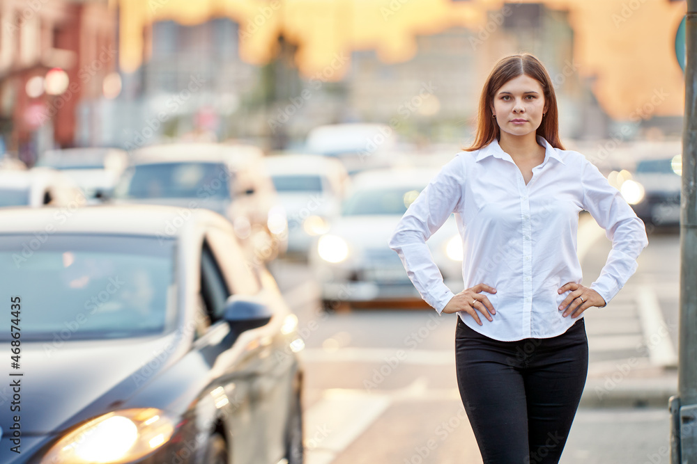 Busy car traffic in center of big city in evening, young woman stands on road with her hands on hips.