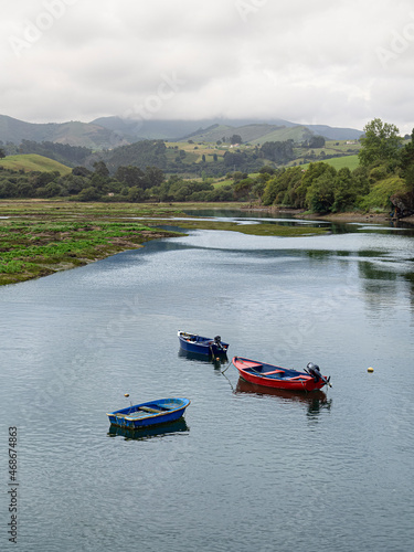 Paisaje de San Vicente de la Barquera con tres barcas en el agua, y el reflejo de las nubes y montañas al fondo, en Cantabria, España, en el verano de 2020.