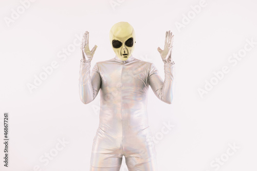 Person dressed in silver suit and green alien mask raising hands in surprise, over white background