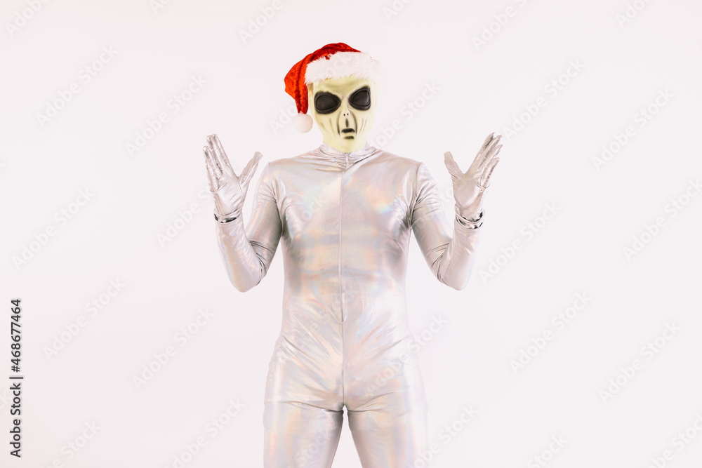 Person dressed in silver suit and green alien mask, with a Christmas cap of Santa Claus, raising his hands in surprise, on white background Person dressed in silver suit and green alien mask, with a Christmas cap of Santa Claus, raising his hands in surprise, on white background
